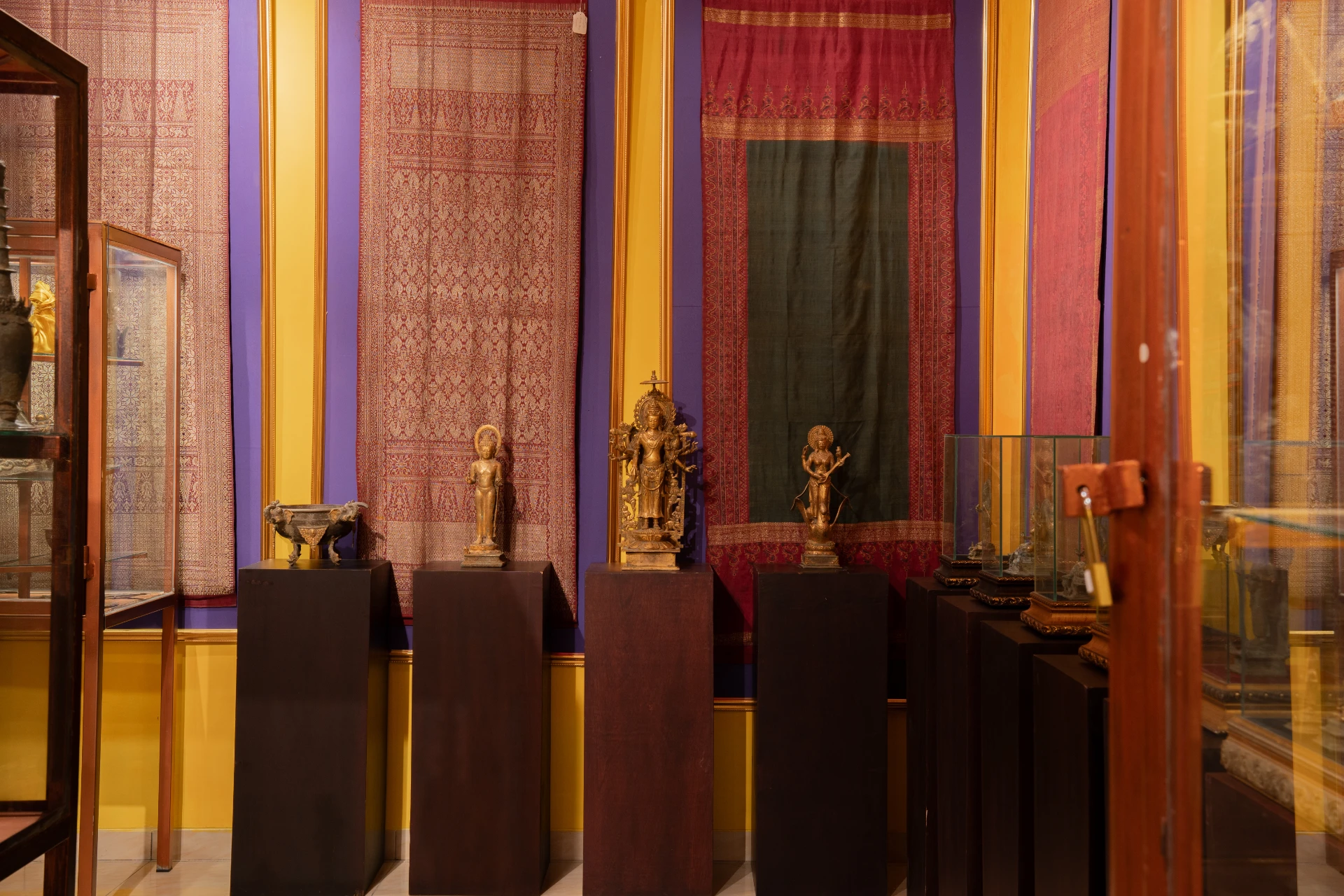 Traditional bronze deity sculptures displayed against tall purple and patterned textile wall hangings, Museum Peter Bloch Collection.