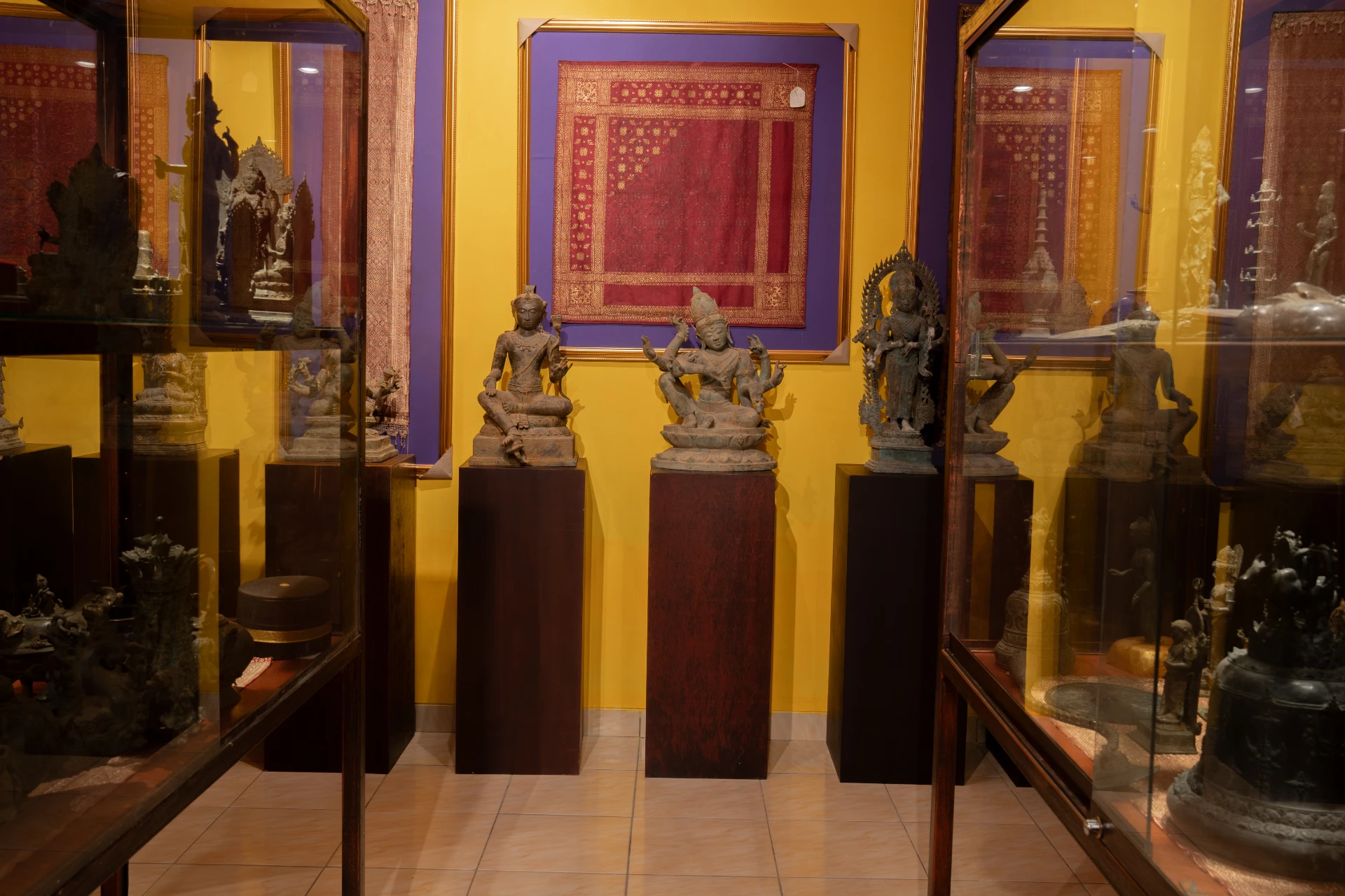 Ancient Hindu-Buddhist bronze statues on dark pedestals with framed traditional textiles, Museum Peter Bloch Collection.